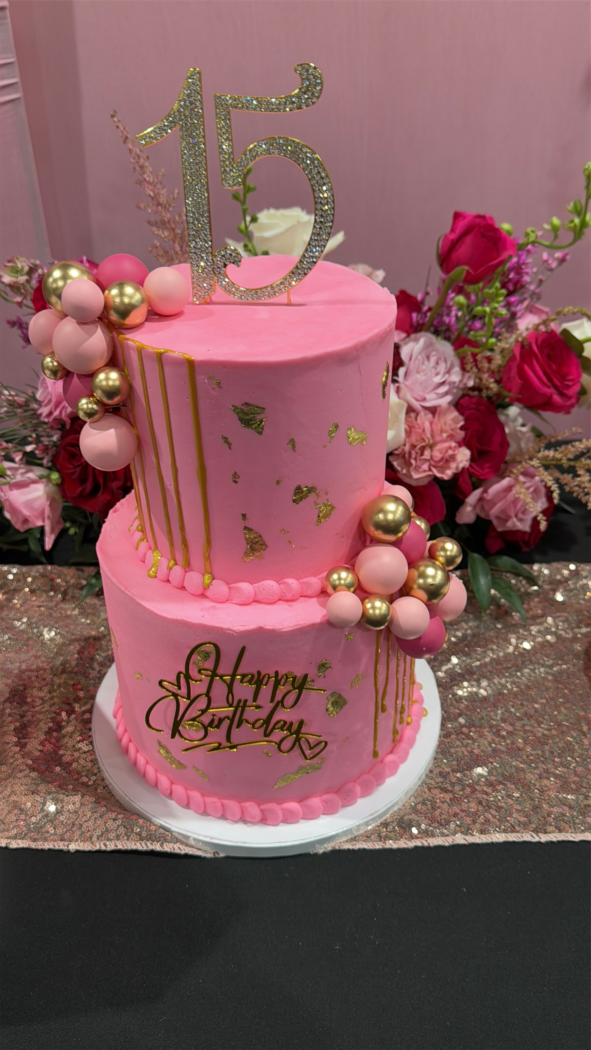 Pink and gold two-tier quinceañera cake with a sparkly 15 topper surrounded by a lush pink and red floral arrangement